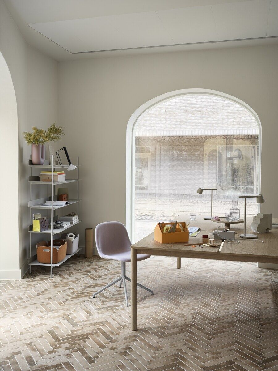 Ein modernes und minimalistisches Home Office mit einem großen Fenster, das viel Tageslicht hereinlässt. Ein Schreibtisch mit diversen Büromaterialien und eine Tragbare Lampe »Piton« auf der Oberfläche. Eine Regalwand mit mehreren Gegenständen und Dekorationen steht im Hintergrund.