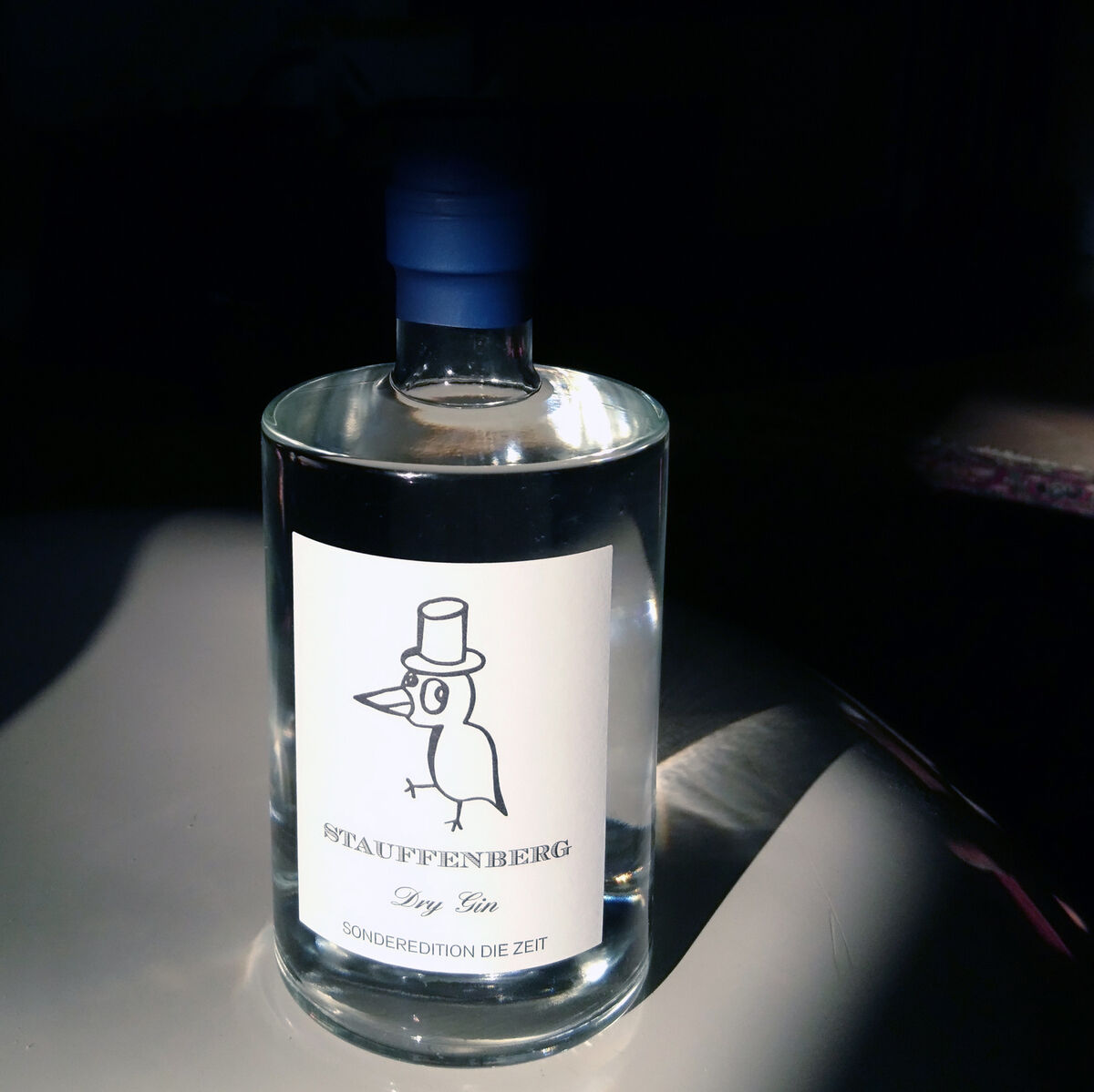 Eine Flasche von Stauffenberg Gin ZEIT Sonderedition, mit einem eleganten Etikett, das einen illustrierten Vogel mit Hut zeigt. Die Flasche steht in einem Lichtstrahl auf einem Tisch in einem ansonsten dunklen Raum.