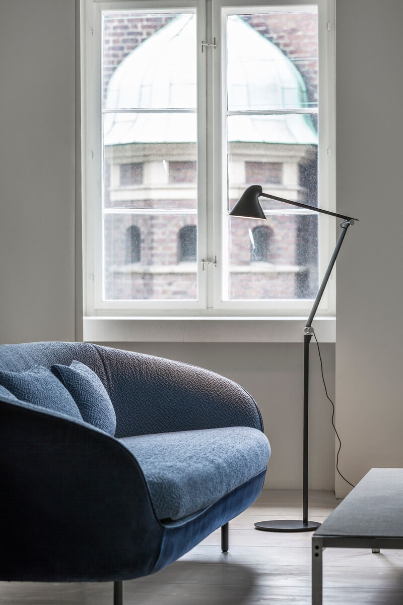 Moderne schwarze Stehleuchte »NJP« neben einem blauen Sofa vor einem Fenster in einem hellen Raum mit Blick auf ein Gebäude im Hintergrund.