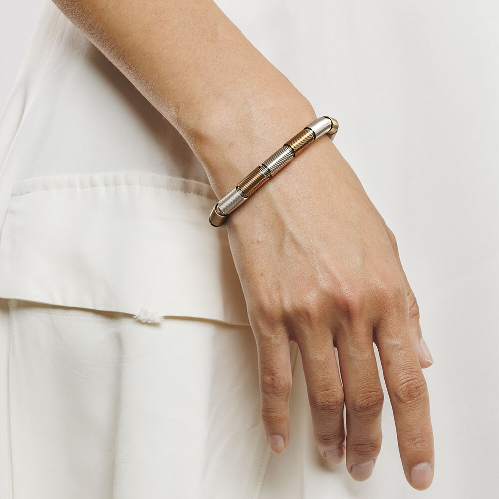 Ein elegantes Armband aus Metall mit Silber- und Goldakzenten, getragen am Handgelenk einer Person mit heller Haut, die einen cremefarbenen Ärmel trägt.