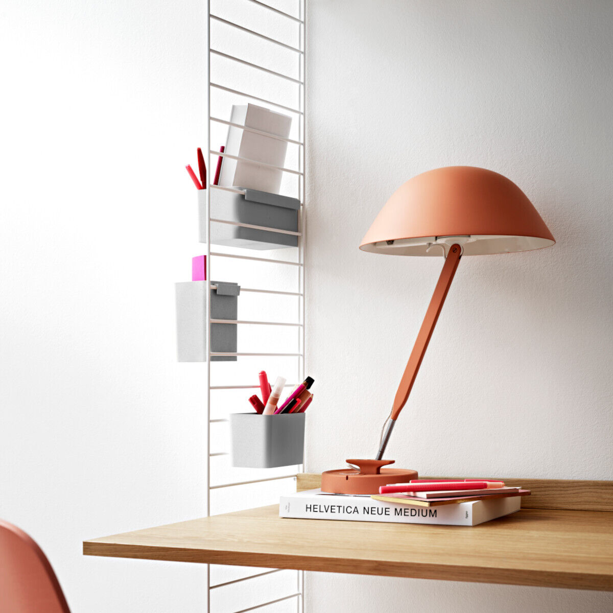 Ein moderner Arbeitsplatz mit weißer Wand, schwebenden Regalböden und einer korallenfarbenen Tischlampe. Auf dem Holztisch liegen Notizbücher und Schreibutensilien.