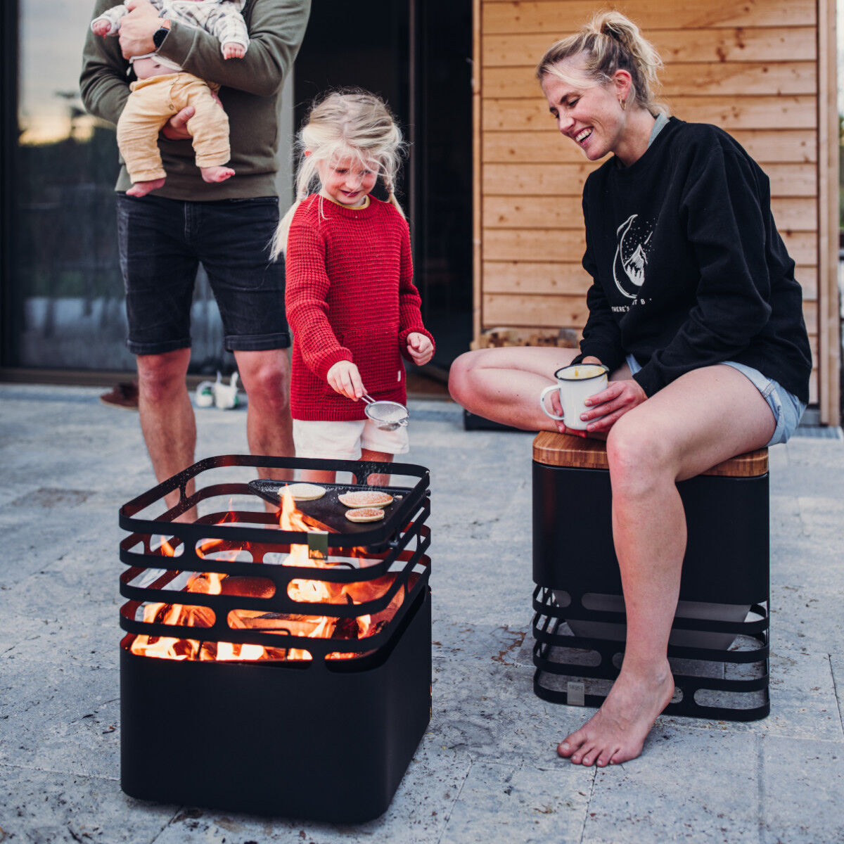 Eine Familie mit zwei kleinen Kindern sitzt draußen auf einer Terrasse neben einer modernen, schwarzen »CUBE« Plancha-Feuerstelle. Der Vater hält ein Baby im Arm, während die Mutter und das andere Kind auf einem Holzhocker sitzen. Es ist ein gemütlicher Abend mit einem warmen Feuer.