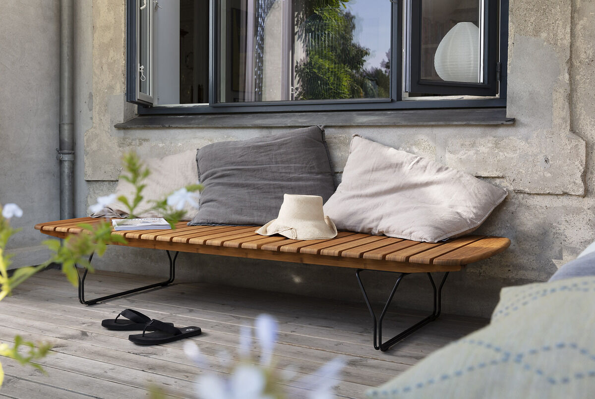 Eine stilvolle Liege namens MOLO aus Holz steht auf einer Terrasse vor einem Haus. Die Liege ist mit hellgrauen und grauen Kissen dekoriert. Neben der Liege liegt ein Strohhut, während im Hintergrund ein Pflanzenarrangement zu sehen ist.