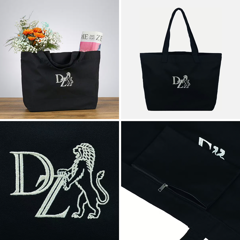 Schwarze Canvas-Tasche mit weißen gesticktem DZ-Logo und Löwe, mit langen Henkeln, gefüllt mit Blumen und Zeitung, Detailansichten von Logo und Reißverschlussfach