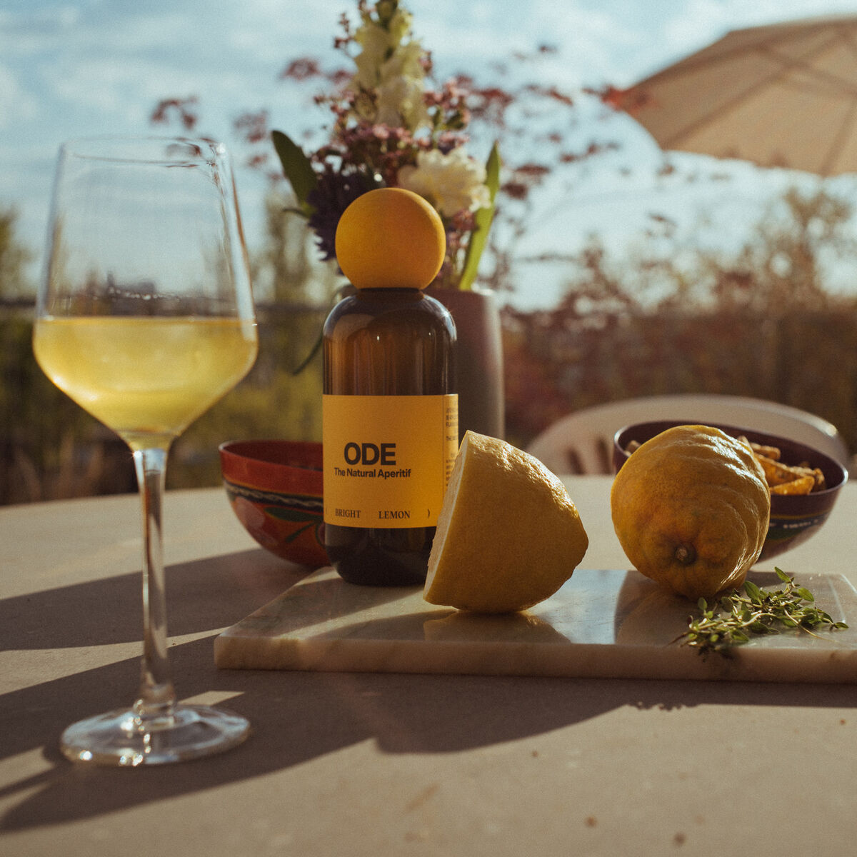Ein Aperitif mit Zitronengeschmack namens 'Bright Lemon', arrangiert auf einem Tisch im Freien mit einem Weinglas, Zitronen und einem Sonnenschirm im Hintergrund.