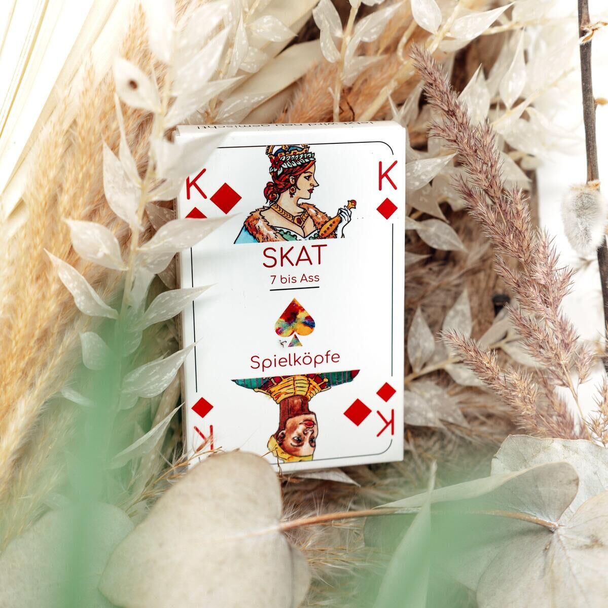 Skatspielkarten-Set mit König, Dame und Bube in klassischem Design, vor einem Hintergrund mit getrockneten Pflanzen und Naturdekor.