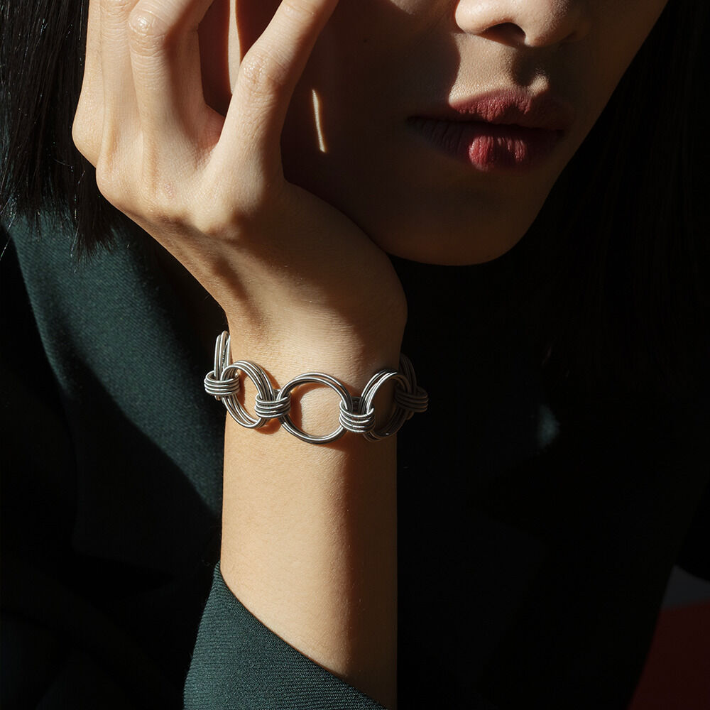 Eine Person trägt ein silbernes Armband mit großen runden Gliedern, das am Handgelenk liegt. Der Hintergrund ist dunkel, und das Gesicht der Person ist teilweise sichtbar.