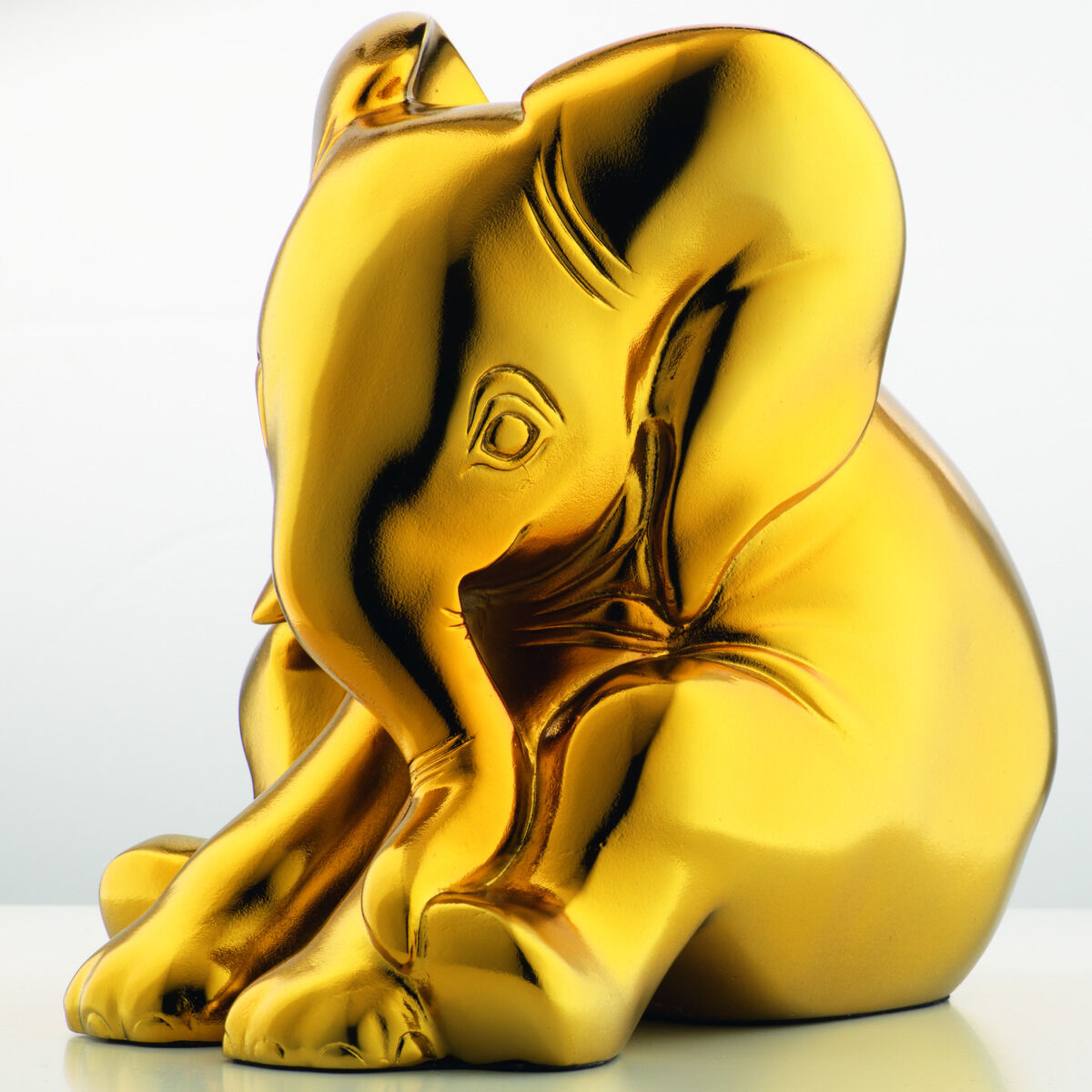 Eine goldene Elefantenskulptur, die aus Kunstguss gefertigt wurde. Der Elefant ist detailliert gestaltet und sitzt aufrecht. Das Bild zeigt die Skulptur vor einem hellen, neutralen Hintergrund.