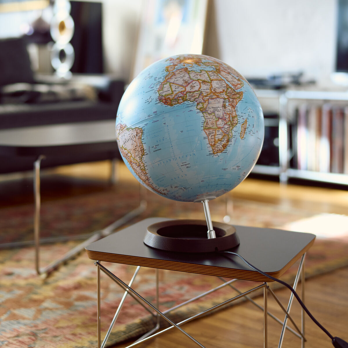 Ein klassischer Globus von National Geographic steht auf einem Tisch in einem Wohnzimmer. Der Globus zeigt geografische Details mit einem Schwerpunkt auf Afrika unter einem blauen Himmel. Die Umgebung ist modern mit einem Teppich und einem Bücherregal im Hintergrund. Das Bild vermittelt ein Gefühl von Abenteuer und Wissenserweiterung.