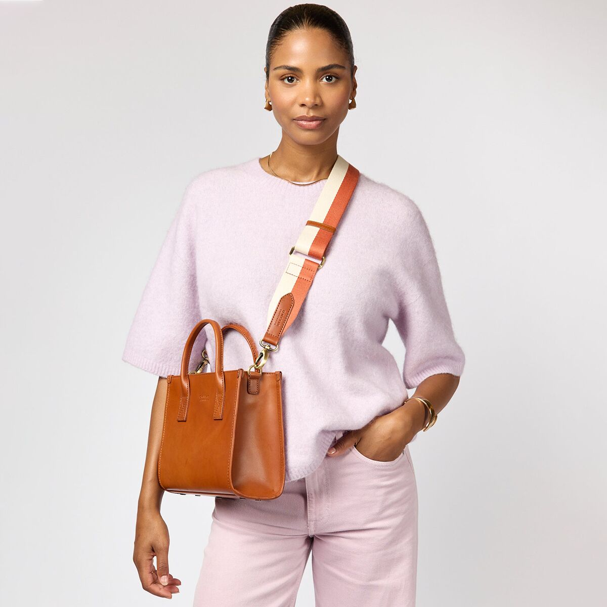 Frau mit hellbrauner Ledertasche »Jackie Midi« mit beigen und goldenen Details, über der Schulter getragen. Die Frau trägt ein helllila Pullover und eine pastellfarbene Hose vor neutralem Hintergrund. Modernes, minimalistisches Modeaccessoire für den Alltag.