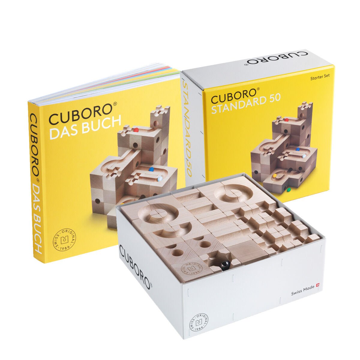 Zu sehen ist das CUBORO Set »STANDARD 50 & DAS BUCH« von Cuboro. Die Verpackung und das geöffnete Set zeigen naturbelassene Holzwürfel für eine Kugelbahn. Daneben steht das dazugehörige Buch mit gelbem Cover, auf dem Kugelbahnbeispiele abgebildet sind.