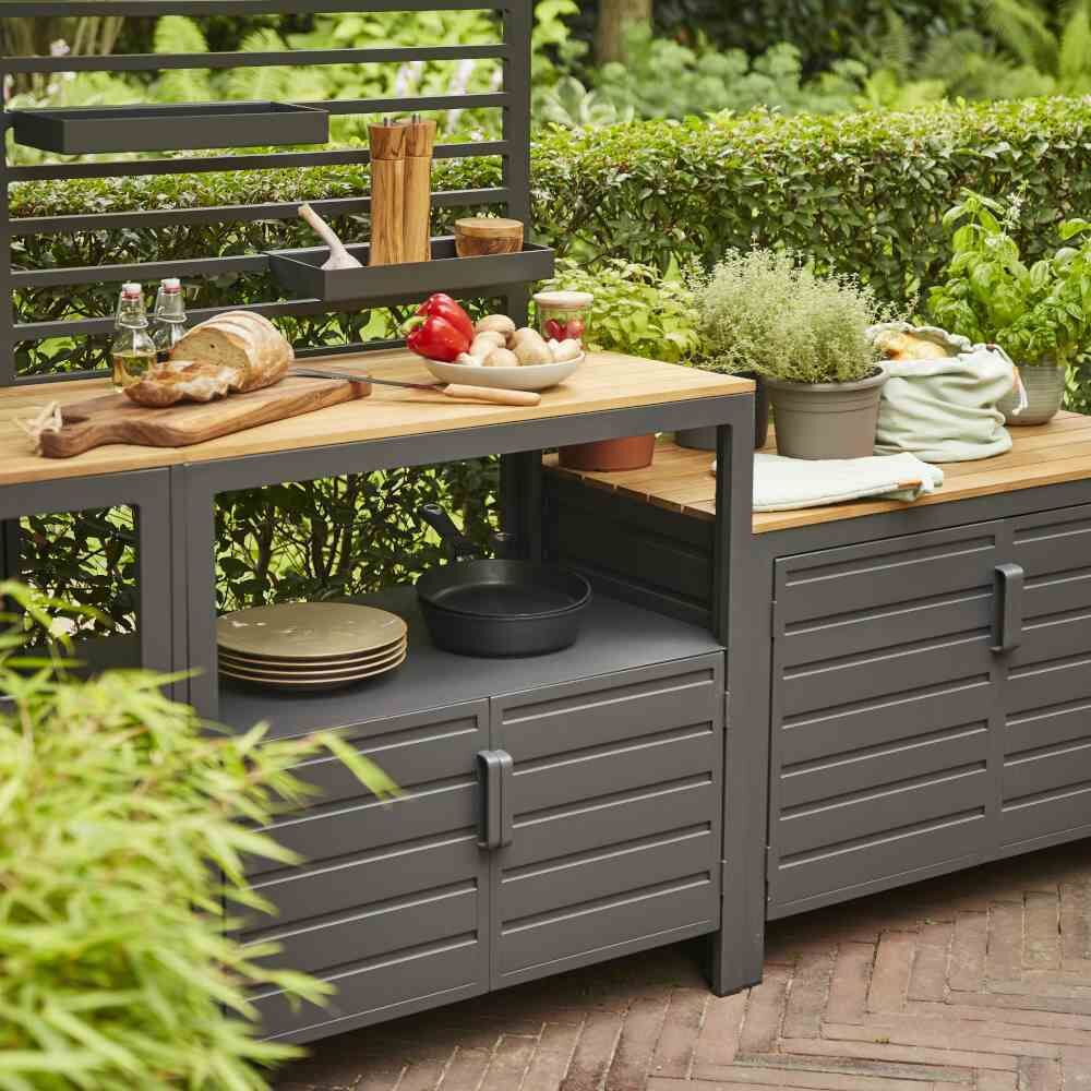 Moderner Outdoor-Küchenschrank 'Sierra' mit grauer Metallstruktur und Holzarbeitsplatte in einem üppig grünen Garten. Auf der Arbeitsplatte befinden sich Küchenutensilien und Gewürzpflanzen.