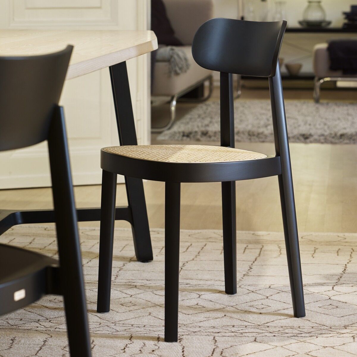 Ein eleganter Stuhl von Thonet, Modell »118«, in einem modernen Esszimmer. Der Stuhl hat ein schwarzes Gestell und eine beige Sitzfläche. Im Hintergrund sind weitere Möbel wie ein Tisch und ein Sofa zu sehen.