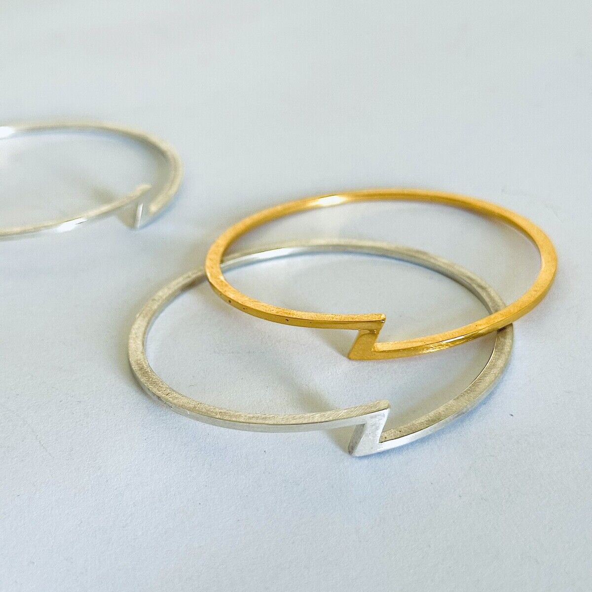 Zwei filigrane Armreifen liegen auf hellem Untergrund. Einer ist aus gebürstetem Silber, der andere vergoldet. Beide zeigen ein modernes, offenes Design mit einer charakteristischen, spitzen Aussparung. Die Schmuckstücke wirken minimalistisch und elegant.