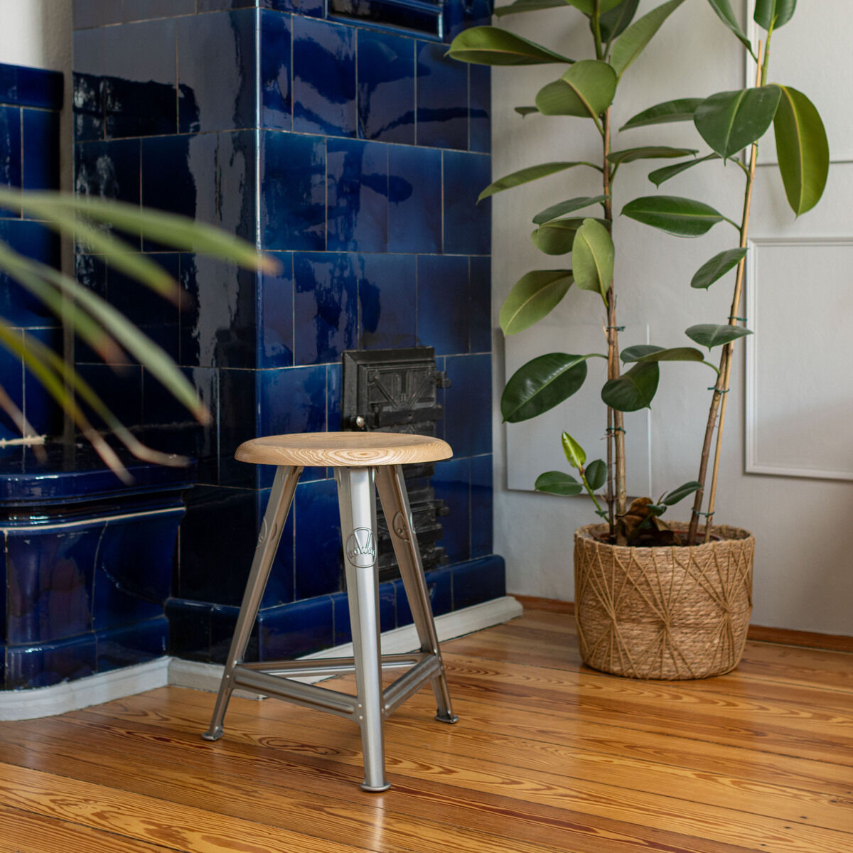 Ein Industriehocker aus blankem Stahl mit runder Esche-Sitzfläche steht auf einem Holzboden. Im Hintergrund sind ein blauer Kachelofen und eine große Grünpflanze in einem geflochtenen Übertopf zu sehen. Die Szene vermittelt ein Gefühl von modernem und gemütlichem Wohnstil.