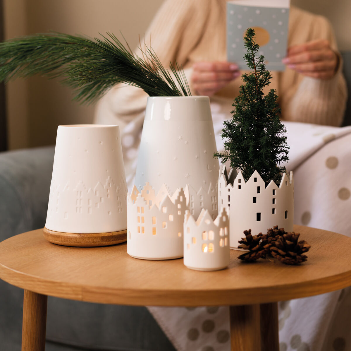 Ein Holztisch mit mehreren Windlichtern in Form von weißen Häusern, darunter einige mit LED-Kerzen, und einer kleinen grünen Tannenbaumdekoration. Im Hintergrund sitzt eine Person in einem gemütlichen Wohnzimmer und liest eine Karte.