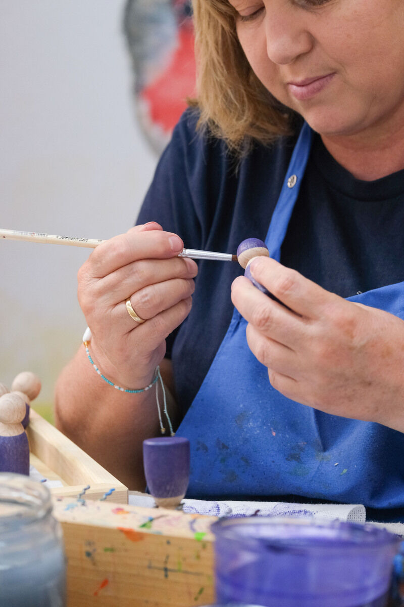 Frau bemalt handgefertigte Holzfiguren in Blau und Lila, die zum Regenbogen Holzspielzeug gehören, mit einem Pinsel. Im Hintergrund sind weitere Holzbausteine und Malutensilien zu sehen.