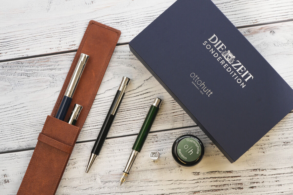 ZEIT-Sonderedition Kugelschreiber »Design04«, bestehend aus einem Füllfederhalter und einem Kugelschreiber in einer eleganten braunen Ledertasche, präsentiert neben einer hochwertigen Geschenkbox und einem Tintenfass auf einem weißen, strukturierten Holztisch.