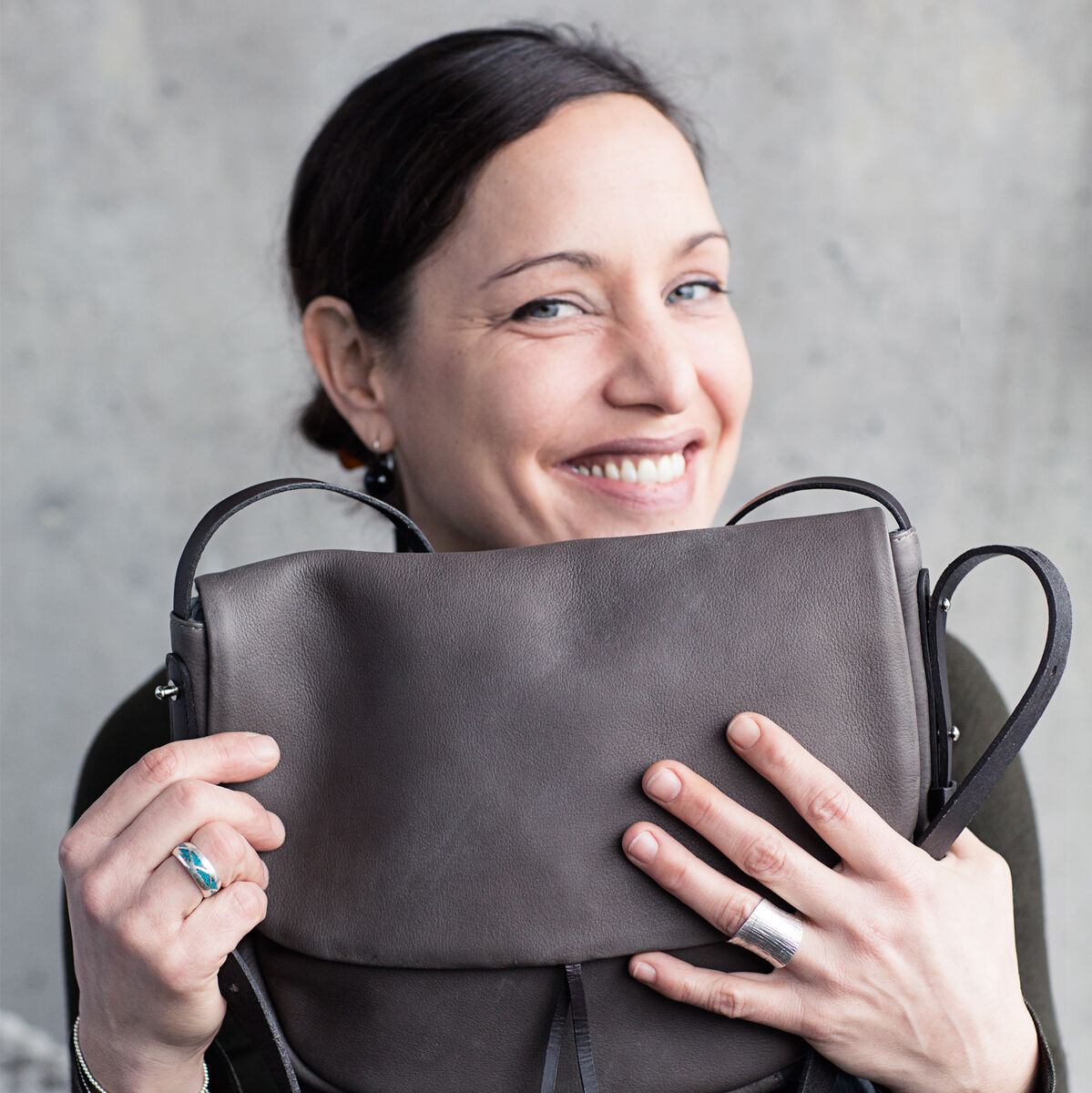 Eine lächelnde Frau hält eine braune Schultertasche »Slingbag M« aus Leder. Die Tasche hat einen einfachen, eleganten Stil mit schwarzen Riemen und einer dunkelbraunen Oberfläche.
