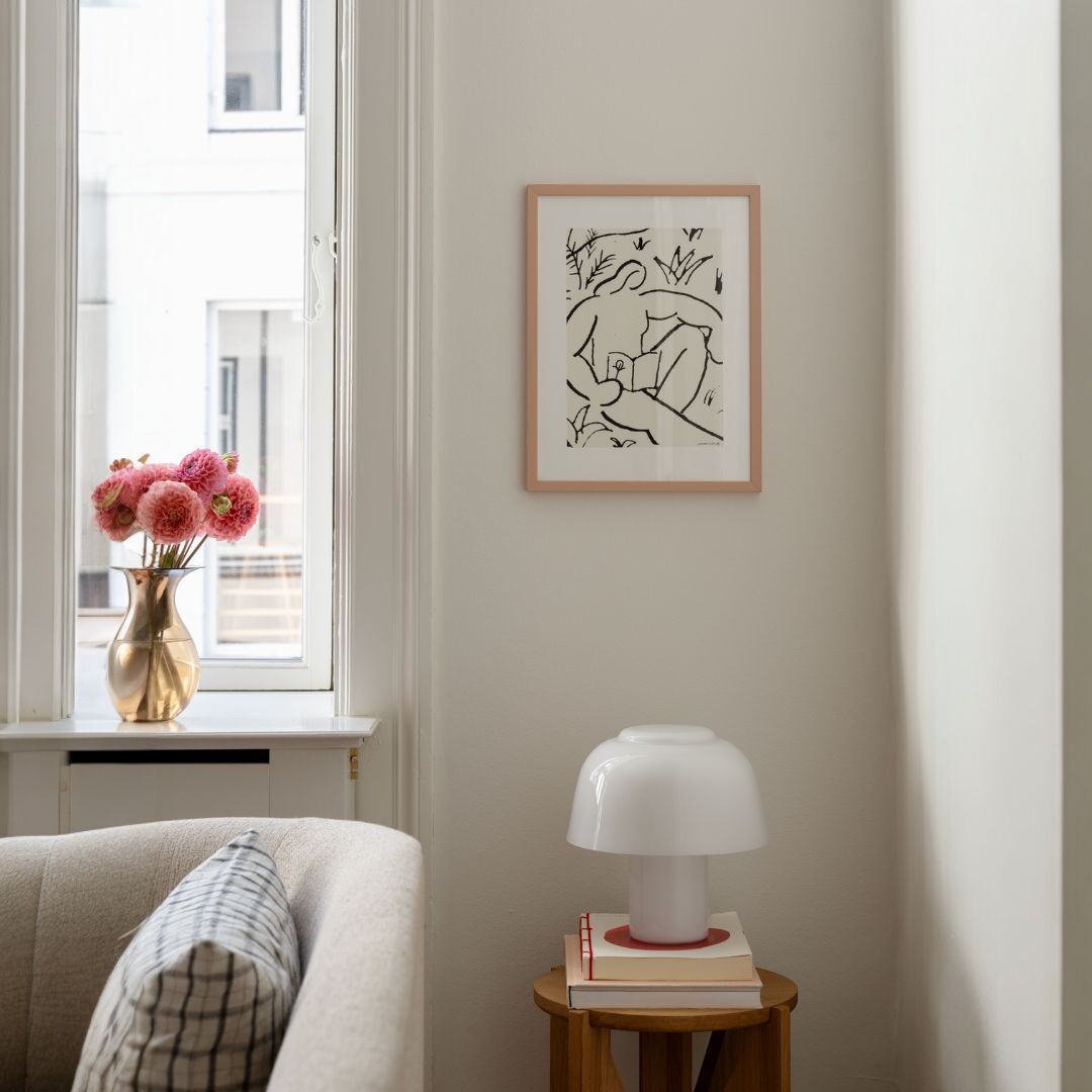 Gemütlicher Wohnraum mit einem minimalistischen gerahmten Kunstwerk an der Wand. Eine Vase mit Blumen steht auf der Fensterbank, daneben ein Sofa und ein Tisch mit einer Lampe und Büchern. Ruhige, sanfte Farben bestimmen das Bild.