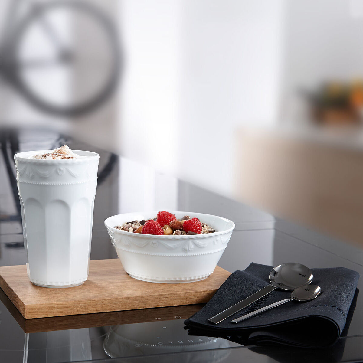 Ein Muesli-Gourmet-Set mit weißer Tasse und Schale, gefüllt mit Joghurt und roten Früchten, steht auf einem hölzernen Brett auf einem modernen schwarzen Tisch, begleitet von glänzendem Besteck.