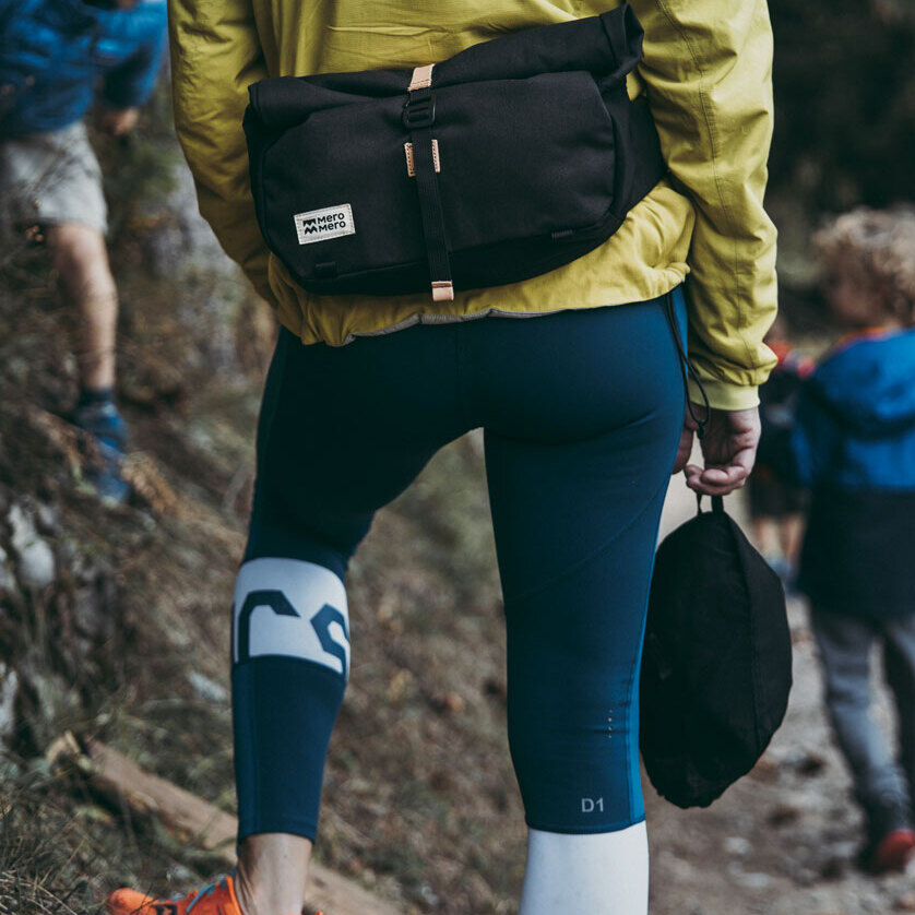 Eine Person trägt eine funktionelle schwarze Tasche »Piha« beim Wandern in einem bewaldeten Gebiet, begleitet von zwei Kindern in blauen Jacken.