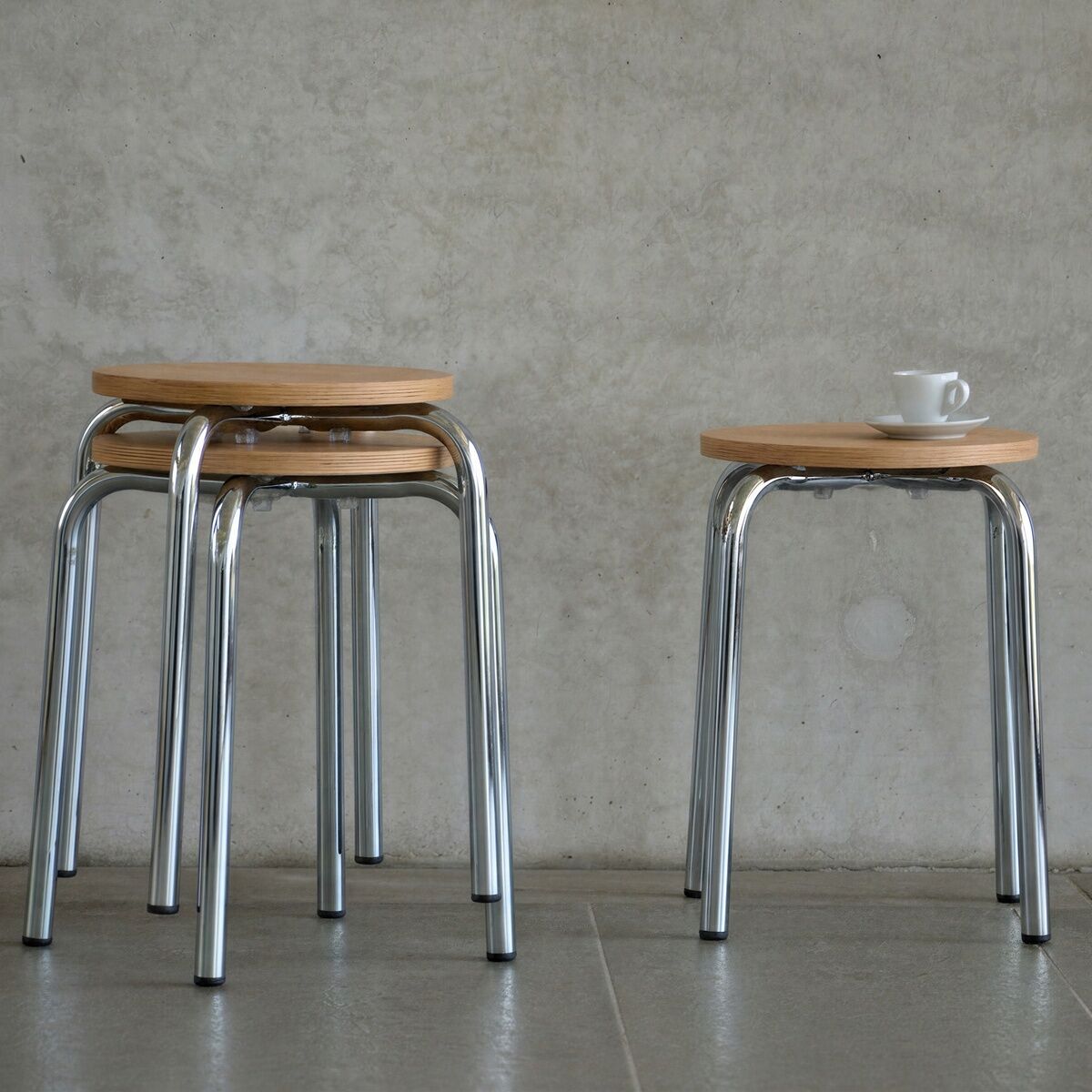 Zwei stapelbare Hocker »PARIS« von Jan Kurtz mit runden Holzsitzen und glänzenden Chrombeinen stehen vor einer grauen Betonwand. Ein Hocker trägt eine weiße Espressotasse, die anderen sind platzsparend ineinandergestellt. Die Szene wirkt modern und schlicht.