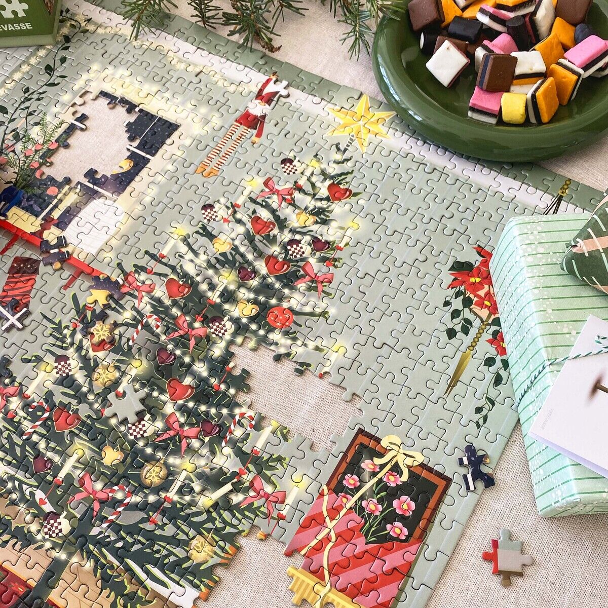 Das Bild zeigt das 1000-Teile Puzzle »Faule Tage« von ViSSEVASSE, fast fertig gelegt. Zu sehen sind ein festlich geschmückter Weihnachtsbaum, Geschenke, Dekoration und einige fehlende Teile. Daneben liegt eine Pralinenschale und ein weiteres Geschenkpaket.