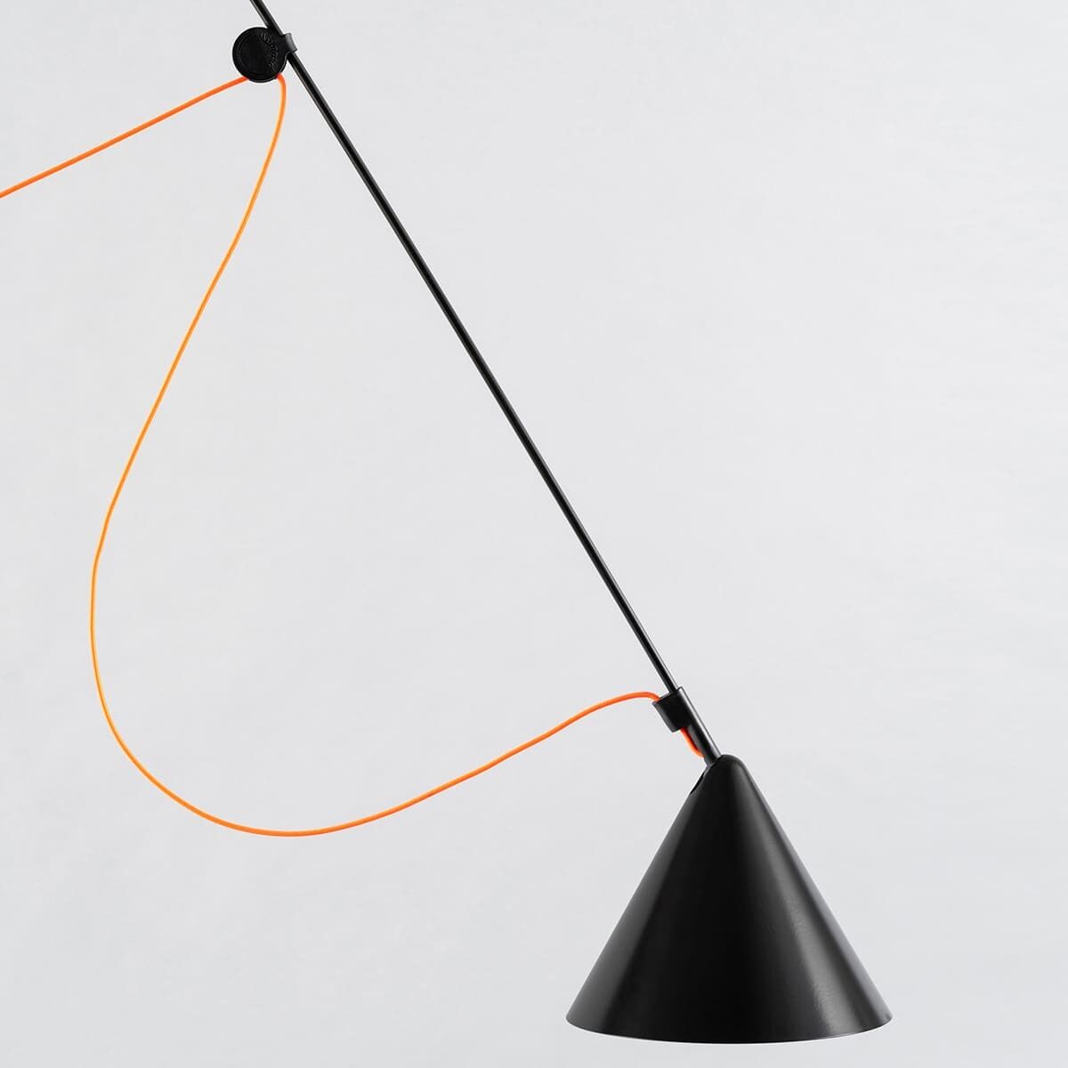 Das Bild zeigt eine moderne Stehlampe mit dem Namen »AYNO«. Die Lampe hat ein schwarzes, konisches Lampenschirm und eine dünne Stange aus schwarzem Metall. Ein auffälliges orangefarbenes Kabel verläuft entlang der Stange.