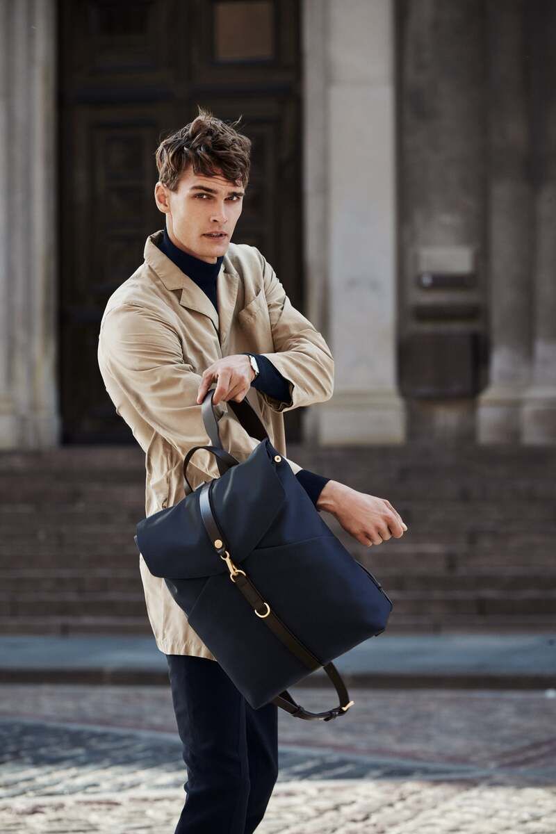 Ein Mann in beiger Jacke trägt einen Rucksack von MISMO, der stilvoll, elegant und praktisch ist. Der Rucksack ist schwarz mit braunen Lederriemen, gut geeignet für Reisen und den täglichen Gebrauch in der Stadt.