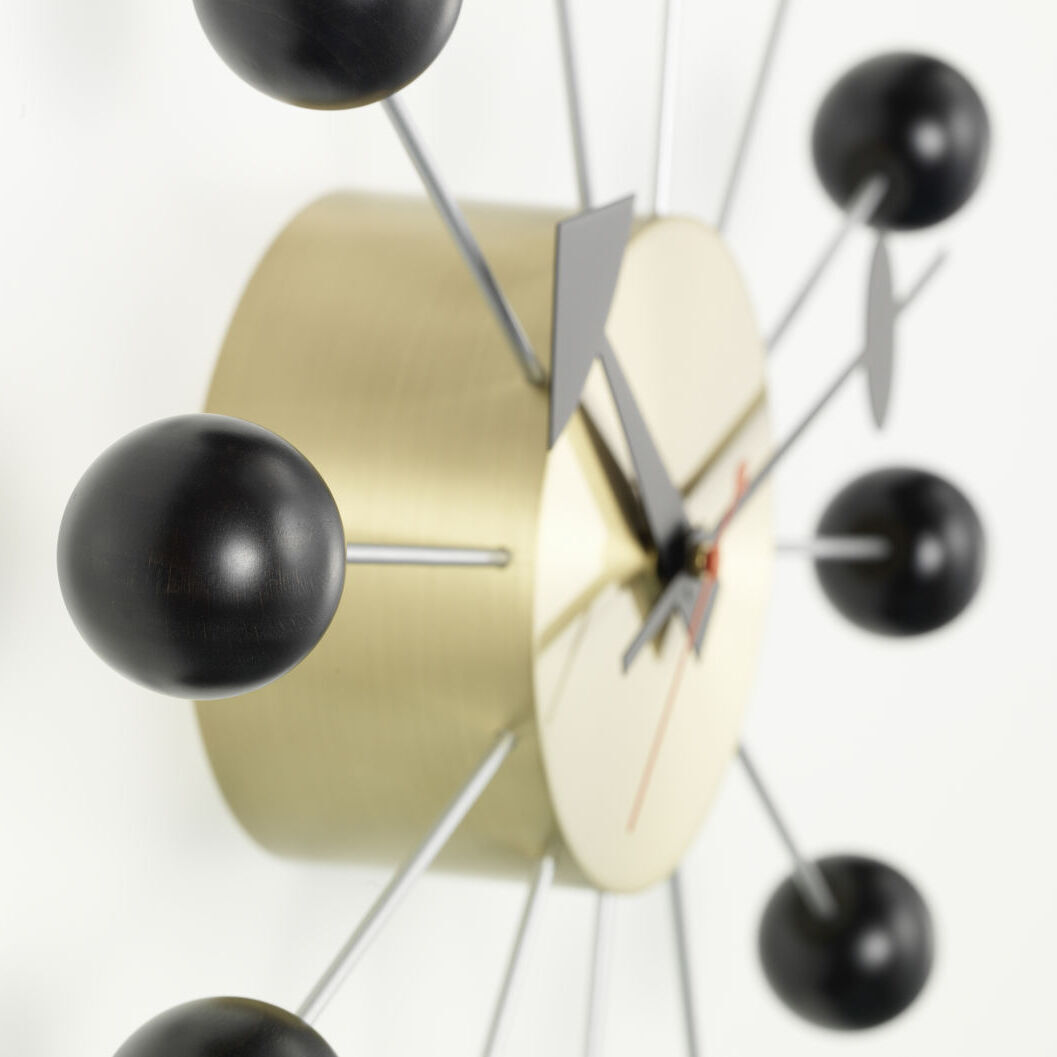 Eine moderne Wanduhr namens 'Ball Clock' mit einer goldenen Basis, schwarzen Kugeln an den Enden von silbernen Metallstäben und filigranen Zeigern. Die Uhr hat ein minimalistisches und stilvolles Design.