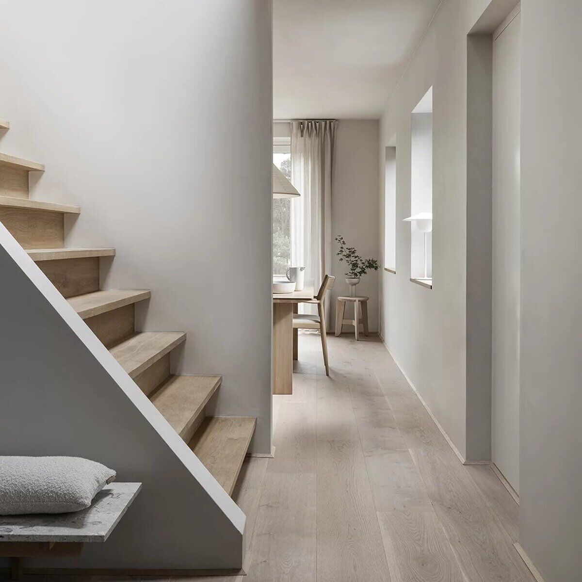 Moderner Sitzhocker aus hellem Holz steht im minimalistischen Flur eines Hauses mit Holztreppe, hellem Boden und viel Tageslicht. Im Hintergrund befindet sich ein Raum mit Tisch und Stuhl. Der Stil ist skandinavisch, schlicht und einladend.