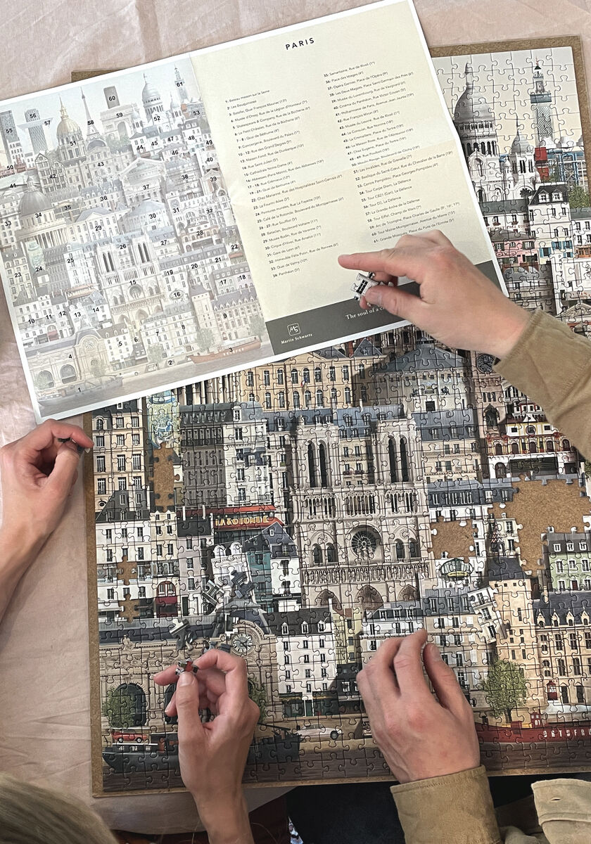 Puzzle mit 1000 Teilen zeigt die Stadt Paris mit markanten Sehenswürdigkeiten und Details. Hände bauen das Puzzle zusammen. Beige, grau, blau, weiße und rote Farbtöne sind vorherrschend. Eine Anleitung ist ebenfalls abgebildet.