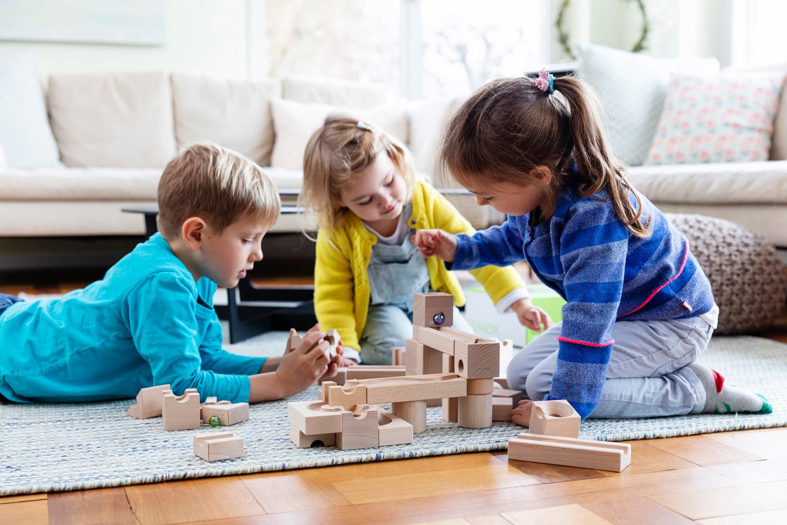 Drei Kinder spielen gemeinsam mit einem Holzbausteinspiel auf dem Boden in einem Wohnzimmer