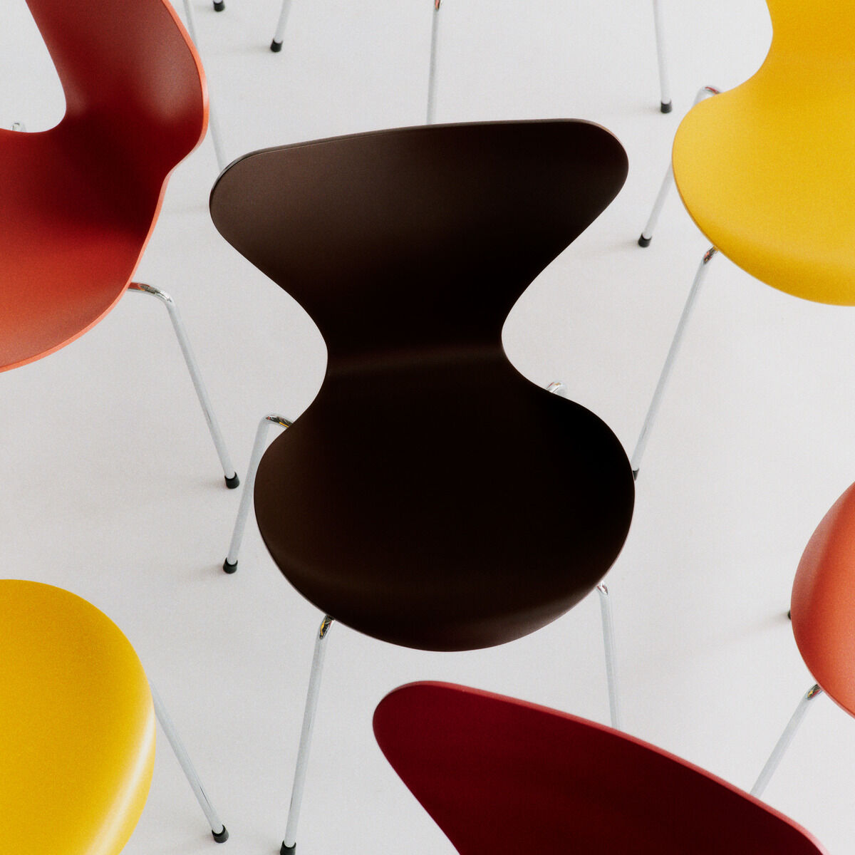 Mehrere Stühle der Serie 7 Edition Verner Panton von Fritz Hansen sind auf hellem Boden arrangiert. Die Stühle haben stilvolle, organisch geformte Sitzschalen in Braun, Gelb, Rot und Orange auf filigranen Chrombeinen.