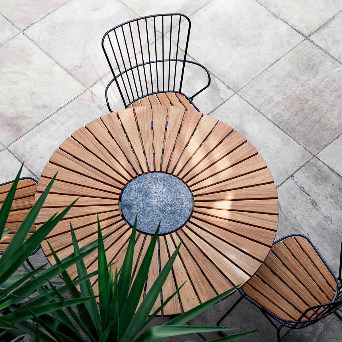 Runder Gartentisch aus Holz und Metall mit zwei passenden Stühlen, auf einem Terrassenboden platziert, umgeben von Pflanzen.