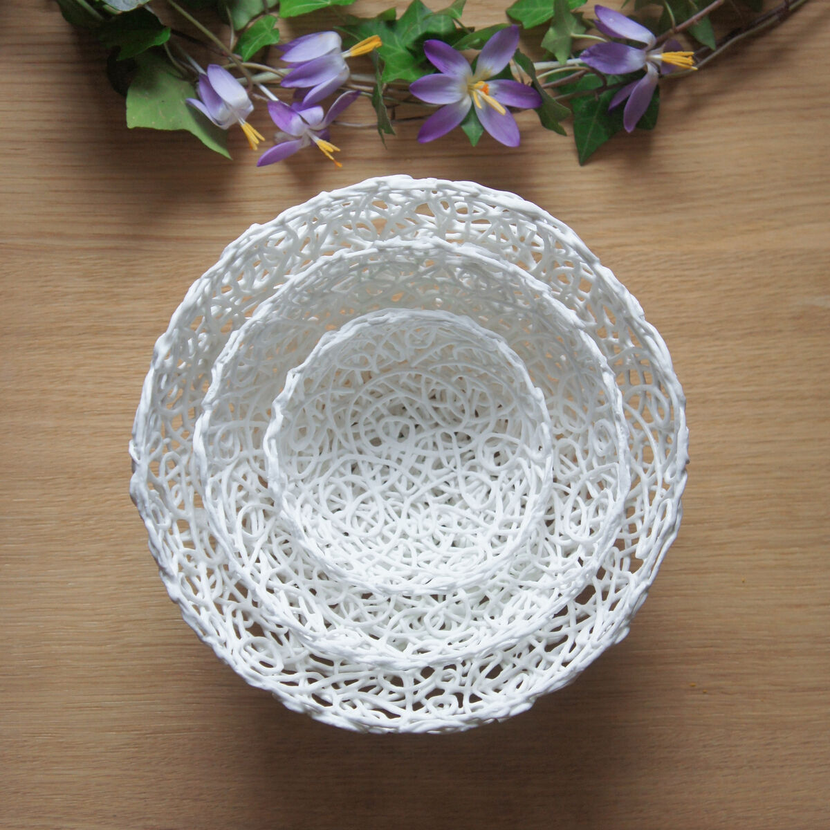 Verschachtelte weiße Porzellanschalen im klöppelartigen Design, umgeben von einer Girlande aus grünen Blättern und lila Blumen, auf einem hölzernen Hintergrund.