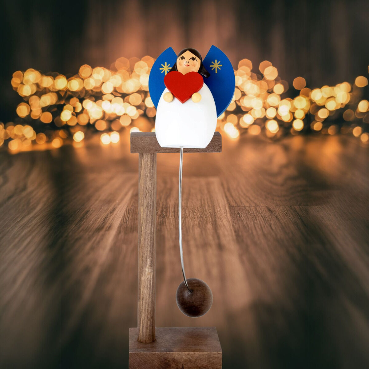 Vor unscharf leuchtenden goldenen Lichterketten steht ein Schaukel-Engel auf dunkelbraunem Holzständer. Die weiße Figur hat blaue Flügel mit goldenen Sternen, hält ein rotes Herz und trägt eine Kugel an einer weißen Schnur als Gegengewicht. Holzfußboden im warmen Licht.