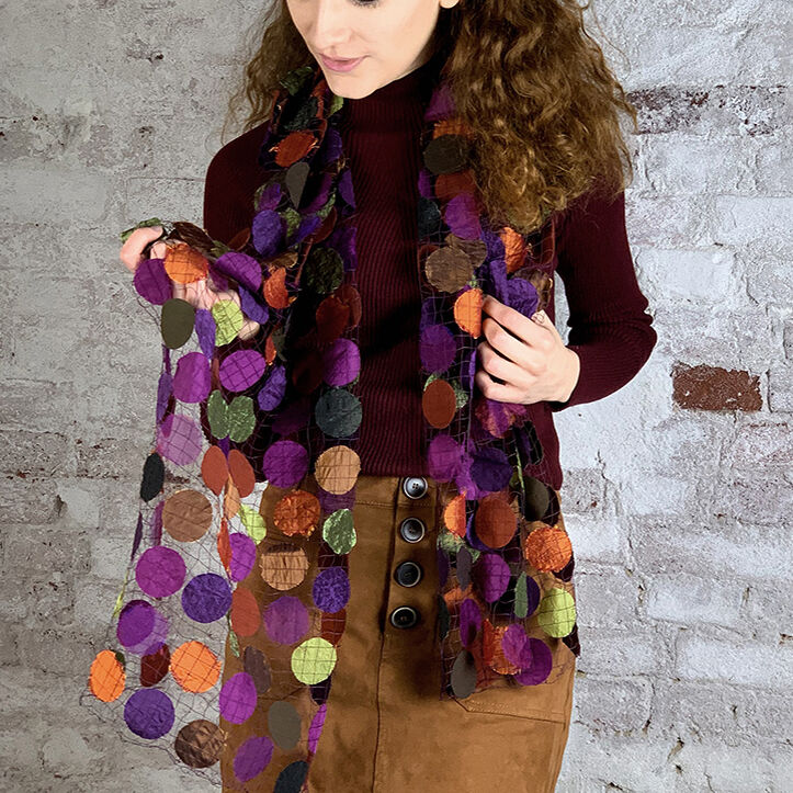 Frau mit lockigem Haar trägt einen transparenten Schal mit Punktemuster in warmen Gewürztönen. Sie hat ein weinrotes Oberteil und einen braunen Rock an und steht vor einer grauen Wand.