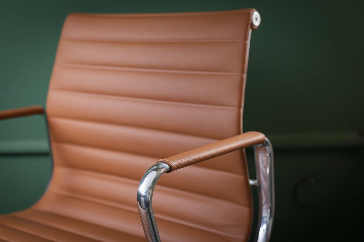 Nahaufnahme eines Vitra Alu Chairs EA 118. Der Bürostuhl hat eine braune Lederpolsterung und polierte Armlehnen aus Metall. Der Hintergrund ist grün.