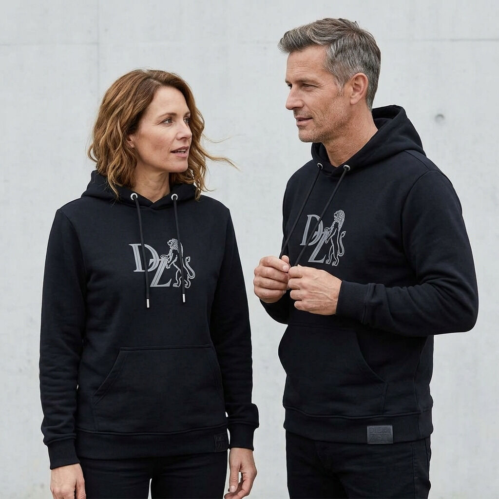 Zwei Personen tragen einen schwarzen ZEIT-Kollektion Hoodie mit auffälligem grauen Schaumprint-Logo und Löwenmotiv auf der Brust. Die Hoodies sind unisex, haben schwarze Kordeln und ein geprägtes Lederlabel am Saum. Der Hintergrund ist hell und schlicht.