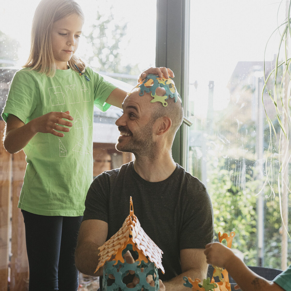 Ein Mann und zwei Kinder spielen mit dem Spiel »Binabo« (60 Teile) an einem Tisch. Ein Kind setzt dem Mann spielerisch Bauteile auf den Kopf. Das Spiel besteht aus farbigen, biegsamen Teilen, die zusammengesetzt werden können, um dreidimensionale Strukturen zu schaffen.