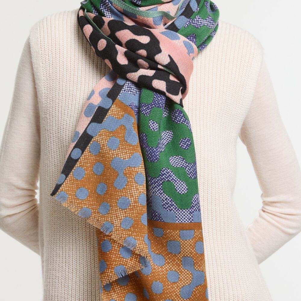 Bunt gemusterter Wollschal in den Farben Grün, Braun, Blau, Schwarz, Rosa und Beige, doppelt um den Hals einer Person gelegt. Der Schal zeigt verschiedene geometrische und abstrakte Muster und wirkt warm, weich sowie modisch. Perfekt geeignet als modisches Accessoire für die Herbst- und Wintersaison.