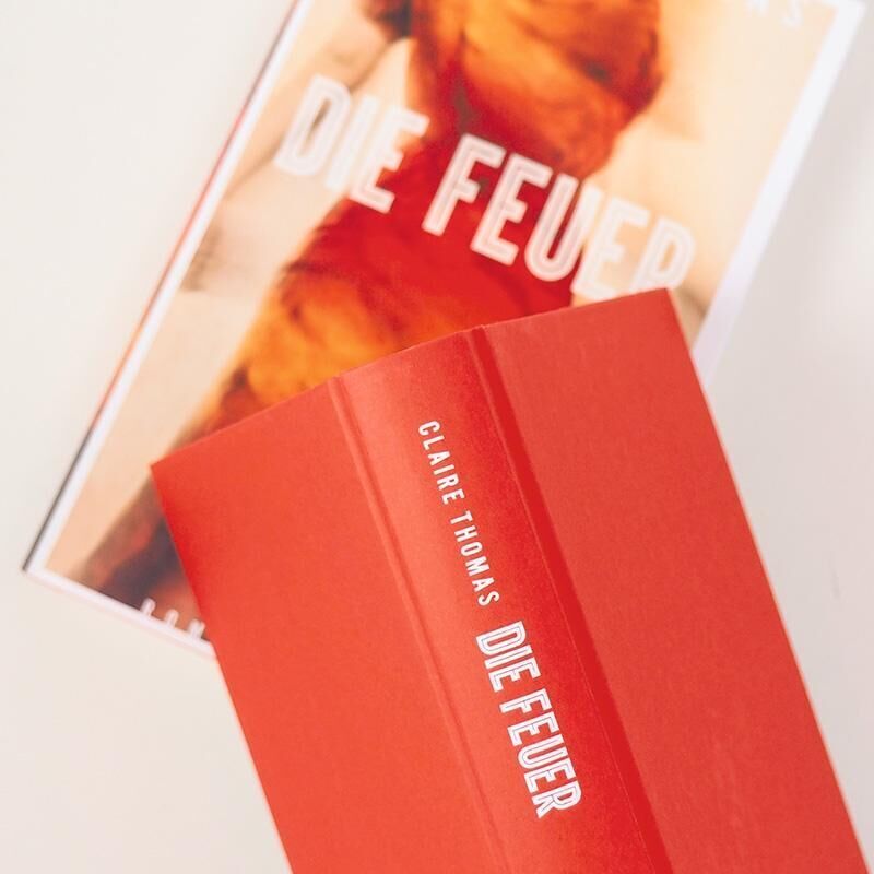 Ein Buch mit dem Titel 'Die Feuer' von Claire Thomas auf einem hellen Hintergrund. Das Cover ist rot mit weißer Schrift und zeigt ein verschwommenes Bild von Feuer.