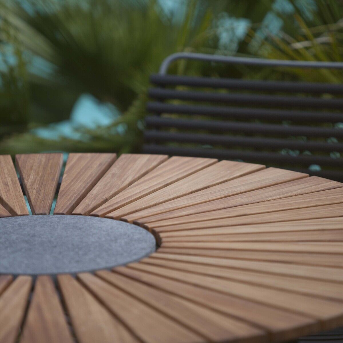 Ein runder Holztisch mit dem Namen 'Circle' steht auf einer Terrasse. Der Tisch hat eine spezielle Gestaltung mit einem zentralen grauen Kreis und strahlenförmig angeordneten Holzleisten. Im Hintergrund sind Pflanzen sichtbar.