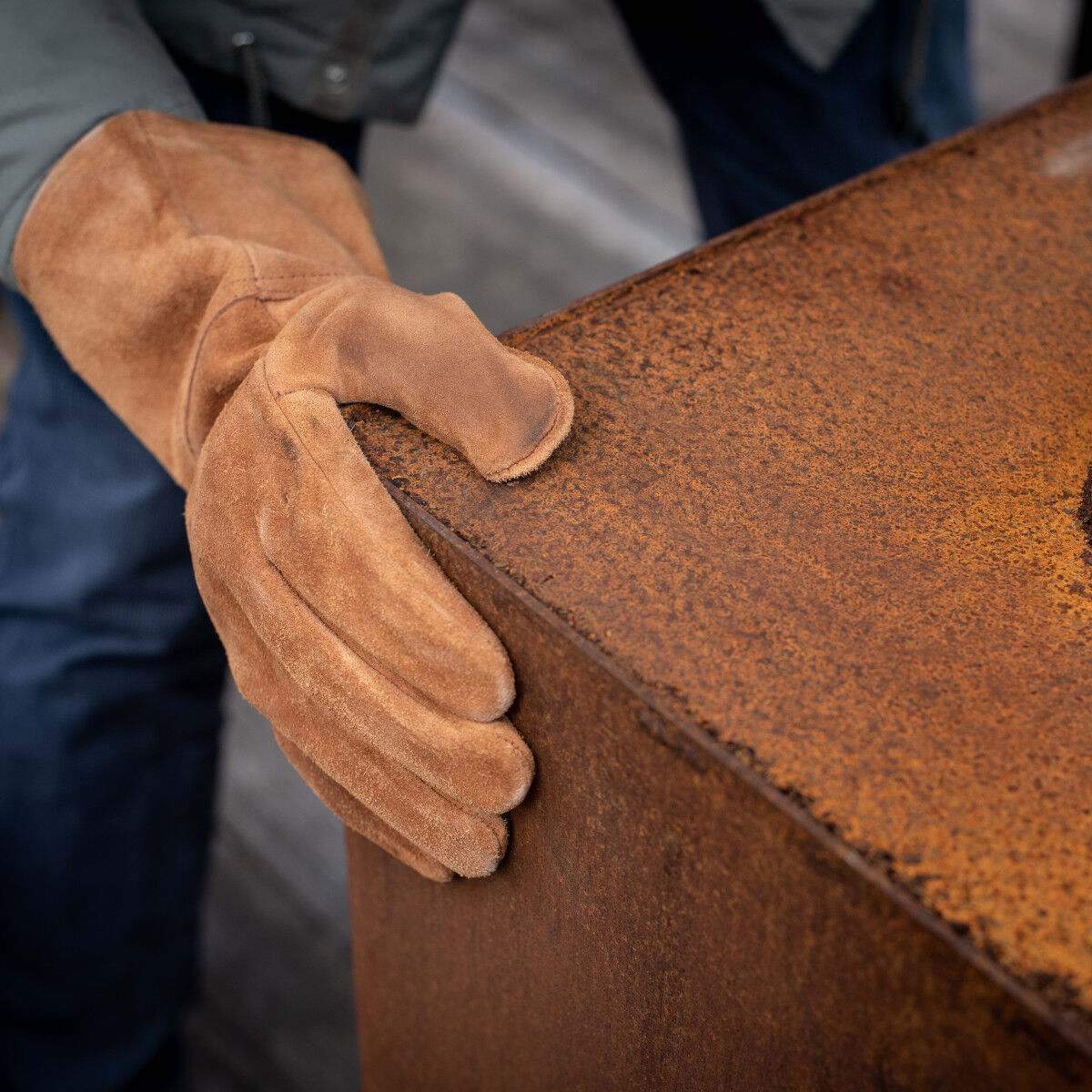 Ein Paar brauner Handschuhe aus Rindleder auf einer rostigen Metalloberfläche. Die Handschuhe werden von einer Person getragen, die für Arbeiten im Freien oder handwerkliche Tätigkeiten gekleidet ist.