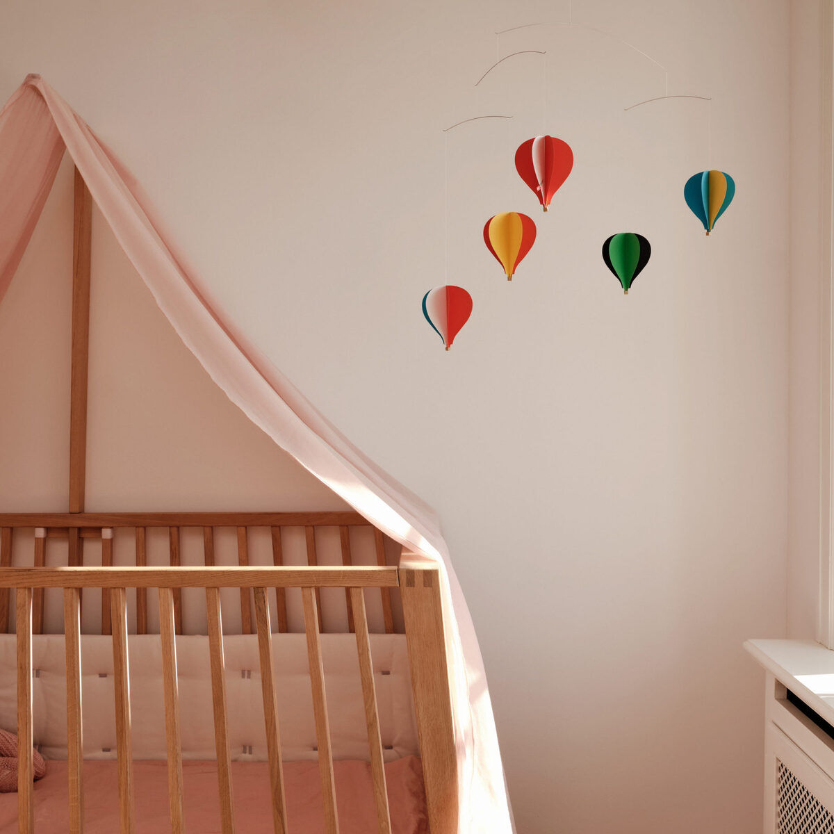 Ein Kinderzimmer mit einem hölzernen Kinderbett und rosa Bettwäsche. Über dem Bett ist eine Wanddekoration mit fünf bunten Heißluftballons in den Farben Rot, Gelb, Grün, Blau und Rosa angebracht.