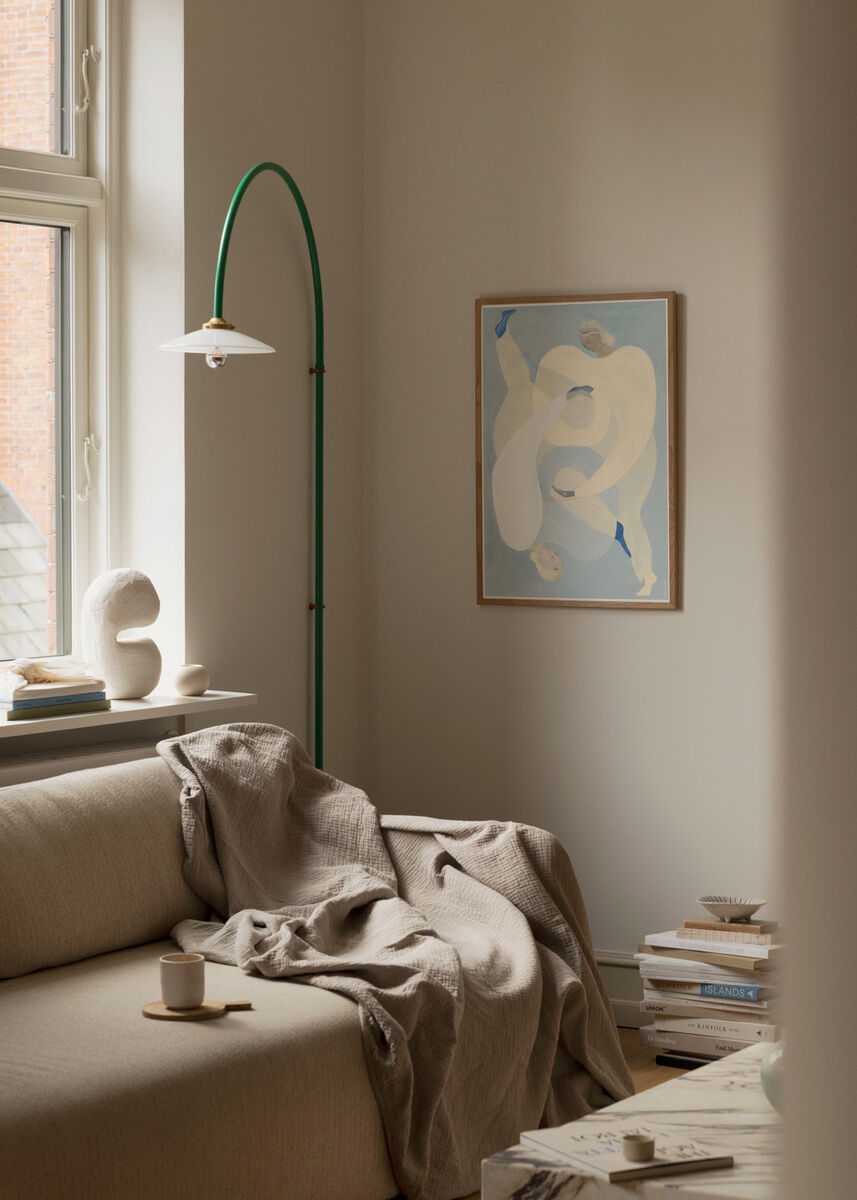 Ein modernes Wohnzimmer mit einem beigen Sofa, auf dem eine Decke liegt. An der Wand hängt ein abstraktes Kunstwerk in Blau- und Beigetönen. Ein grüner Bogenlampenständer steht neben dem Sofa. Ein Fenster lässt weiches Licht in den Raum.