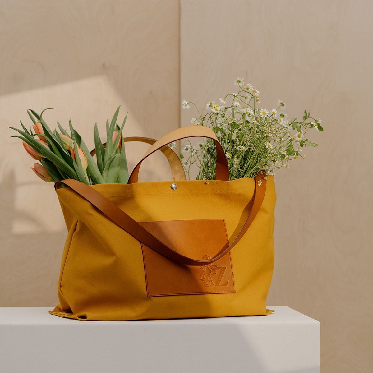 Eine gelbe Jubiläums-Markttasche aus Canvas mit braunen Ledergriffen, gefüllt mit Tulpen und Gräsern, steht auf einem weißen Tisch vor einem hellbeigen Hintergrund.