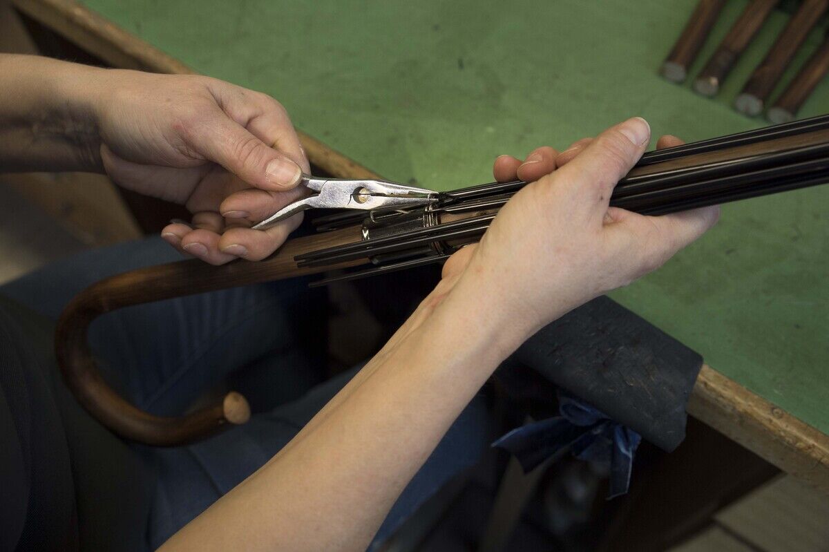 Nahaufnahme von Händen eines Handwerkers, der an einem Regenschirm arbeitet. Der Schirm hat einen klassischen braunen Holzgriff. Die Arbeitsplatte ist grün.